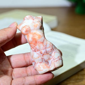 May include: A small, marble-like sculpture of a torso, featuring a white base with red and orange veining. The figurine is held in a hand, with a book and a blurred background.