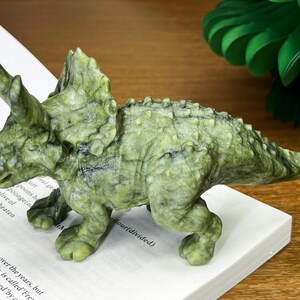 May include: A carved green stone triceratops figurine featuring a textured, mottled surface with dark veining, three distinct horns, and a large frill. The dinosaur is depicted in a standing position with visible skin details and a pointed tail.