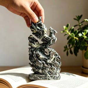 May include: A carved stone sculpture depicting a phoenix rising from flames, featuring a polished black and white marbled texture. The intricate figurine shows the bird with wings spread upward, set upon a solid, flat, rectangular base.