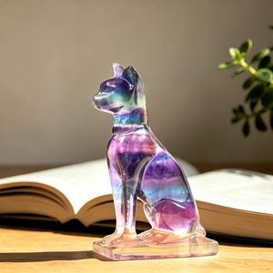 May include: A translucent, carved fluorite figurine in the shape of a seated cat. The stone displays natural colour banding in shades of deep purple, blue, and clear white, with a smooth, polished surface and a rectangular base for stability.