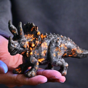 May include: A detailed sculpture of a Triceratops dinosaur. The dark grey stone figure has orange and yellow accents. The dinosaur has three horns, a frill, and a textured body. The sculpture is held in a hand.