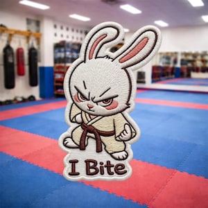 Peut inclure: Écusson brodé représentant un lapin de dessin animé en colère, vêtu d'un uniforme de karaté avec une ceinture marron. Le patch porte l'inscription "I Bite" en police serif. Le lapin a une expression sévère avec des yeux rouges.