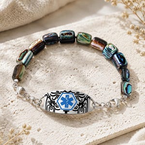 Op de afbeelding: Een medische alarmarmband met iriserende abalone schelpkralen in blauwe, groene en zwarte tinten. De centrale zilverkleurige metalen plaat toont een blauw Star of Life-embleem met een wit symbool van een slang en een staf.