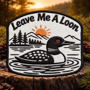 Könnte beinhalten: Gestickter Aufnäher mit dem Text "Leave Me A Loon". Der Aufnäher zeigt einen schwarz-weißen Eistaucher, der im Wasser schwimmt, mit einem Berg- und Sonnendesign im Hintergrund. Der Aufnäher ist schwarz umrandet.