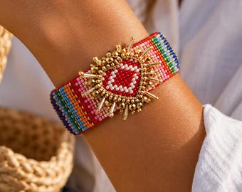 May include: A wide woven beaded bracelet featuring a vibrant rainbow stripe pattern. The center is adorned with a gold-toned heart-shaped emblem surrounded by metallic gold beads and radiating spikes, creating a textured and dimensional focal point.