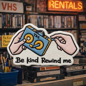 May include: Embroidered patch featuring a blue and yellow cassette tape design held by two hands. The patch reads "Be kind Rewind me" in white text. The background shows a VHS rental store.