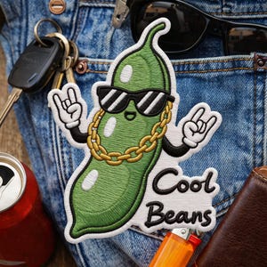 Könnte beinhalten: Gestickter Aufnäher mit einer grünen Bohne, die eine Sonnenbrille und eine goldene Kette trägt, mit dem Text "Cool Beans". Der Aufnäher hat weiße Umrisse und ist vor einem Jeanshintergrund platziert.