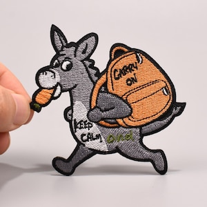 Peut inclure: Écusson brodé représentant un âne gris portant un sac à dos orange avec l'inscription "Carry On". L'âne tient une carotte et l'écusson comprend les mots "Keep Calm and" dans une police stylisée.