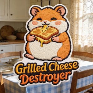 Op de afbeelding: Geborduurde patch met een cartoon hamster met oranje en witte vacht, die een gegrilde kaassandwich eet. De patch heeft de tekst "Grilled Cheese Destroyer" in gele en oranje letters.