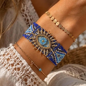 May include: A wide woven bracelet featuring a central gold-toned evil eye charm with blue beads, surrounded by a geometric blue, white, and black pattern. It is accompanied by two thin gold-toned chain bracelets, one with small circular discs.