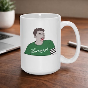 May include: White ceramic mug featuring a cartoon illustration of a person with blood on their mouth and the word "Bazinga!" in green. The person is wearing a green shirt with a star design and black and white stripes on the sleeve.