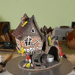 May include: A brown ceramic incense burner shaped like a house with a textured roof, a chimney, and a tree trunk extension. The structure features small yellow accents, red mushroom details, a triangular window, and a central opening for a tea light.