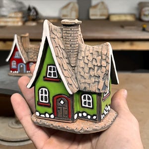 Könnte beinhalten: Eine kleine Keramikhausfigur mit leuchtend grüner Fassade, einem strukturierten braunen Schindeldach, einem zentralen Schornstein und einer dunkelbraunen Bogentür. Das Gebäude hat weiß umrandete Fenster und einen weißen Sockel.