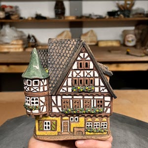 Könnte beinhalten: Detaillierte Keramikhausfigur mit braunem Dach, weißen und braunen Wänden und einem grünen Turm. Das Haus hat mehrere Fenster und dekorative Blumenschmuck. Die Figur wird in einer Hand gehalten.