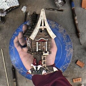 May include: A small, hand-painted ceramic house sculpture featuring a dark brown wood-grain texture, a pointed roof, a central door, and a ladder leading to the entrance. Two red and white spotted mushrooms sit at the base of the structure.