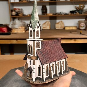 May include: A ceramic church figurine with a steeple and a red tile roof. The church has white walls, brown trim, and arched windows. A cross is visible on the side. The figurine is held in a hand.