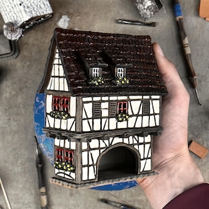May include: A ceramic miniature house featuring a dark brown tiled roof, white walls with black timber framing, red window frames, and yellow flower boxes. The structure includes two levels, a gabled roof with two dormer windows, and an arched opening.