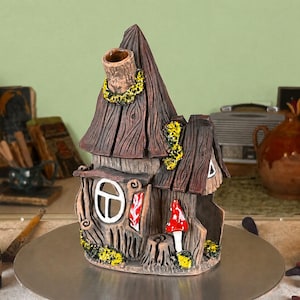 May include: A brown ceramic house-shaped incense burner with a textured wood-grain finish, a tall chimney, a white-framed window, and a red-and-white mushroom accent. The structure features yellow moss-like details and a hollow interior.