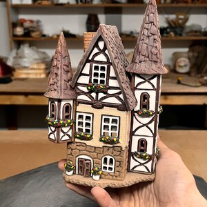 Könnte beinhalten: Ein detailliertes Keramik-Miniaturhaus mit braunem Dach, weißen Wänden und dekorativen Blumenkästen. Das Haus hat zwei Türme und einen Schornstein. Das Äußere ist in Braun- und Beigetönen bemalt, und die Fenster sind schwarz umrandet.