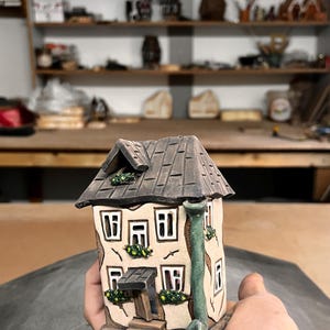 Op de afbeelding: Een keramieken huisfiguur met een bruin dak, beige muren en kleine vierkante ramen. Het huis heeft een groene schoorsteen en decoratieve groene planten. De figuur wordt in een hand gehouden.