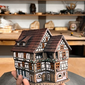 May include: A detailed ceramic miniature house with a brown tile roof, white and brown timber framing, and small orange windows. The house has decorative floral accents and is held in a hand. Other similar houses are visible in the background.