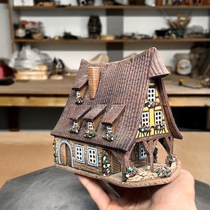 May include: A detailed ceramic miniature house with a brown roof, yellow walls, and decorative floral accents. The house features multiple windows, a chimney, and a small porch, creating a charming, rustic aesthetic.