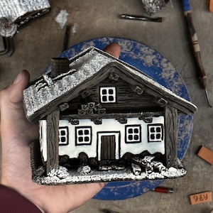 May include: A miniature model of a house with white walls, dark brown wooden beams, and a roof covered in white snow. The structure features a central door, multiple windows with white frames, a chimney, and a small pile of logs near the base.