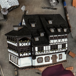 May include: A ceramic miniature house featuring a black shingled roof, white walls with dark brown timber framing, multiple windows, and balconies adorned with small yellow flowers. The structure has a detailed facade with a central wooden door.