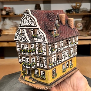Op de afbeelding: Een keramieken miniatuurhuis met een gele basis, witte muren met bruin vakwerk en een donkerrood pannendak. Het bouwwerk bevat meerdere ramen, twee schoorstenen en kleine bloembakken met kleurrijke bloemdetails onder de ramen.