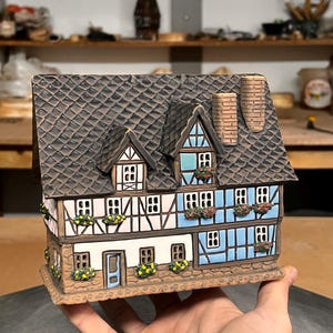 Op de afbeelding: Een keramisch miniatuurhuis met een bruin pannendak, witte en blauwe muren en decoratieve bloembakken. Het huis heeft gedetailleerde ramen, een schoorsteen en een kleine deur, die een rustiek ontwerp laten zien.