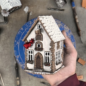 Op de afbeelding: Een miniatuur keramieken huissculptuur met een wit getextureerd dak dat op sneeuw lijkt, muren met een steenpatroon, boogramen en een houten deur. Een kleine rode stoffen zak hangt aan een haakje aan de voorzijde boven de ingang.