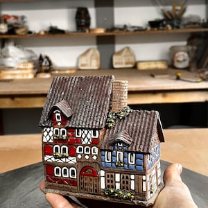 Könnte beinhalten: Ein detailliertes Keramik-Miniaturhaus mit braunem Ziegeldach, roten und blauen Wänden und weißen Fensterrahmen. Das Haus hat einen Schornstein und ist mit kleinen gelben Blumen geschmückt. Das Haus wird in der Hand einer Person gehalten.