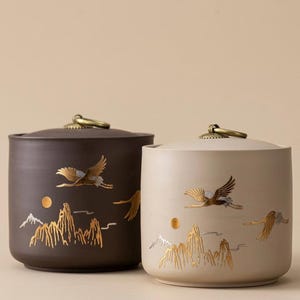 Op de afbeelding: Twee keramische potten met deksels. Eén is donkerbruin, de andere beige. Beide hebben gouden illustraties van vliegende vogels, bergen en een zon. Elke pot heeft een messingkleurige handgreep op het deksel.