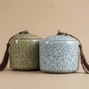 May include: Two ceramic jars with a crackle glaze finish. One is light green, the other is light blue. Each has a bronze-coloured lid with a tassel. The jars are cylindrical with rounded tops, suitable for storage or display.