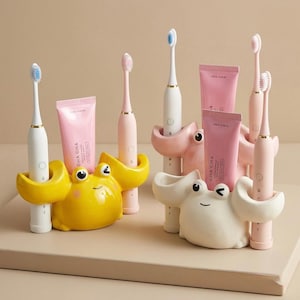 Puede incluir: Una colección de cepillos de dientes y tubos de pasta de dientes exhibidos en soportes con forma de rana. Los soportes son amarillos, rosas y blancos. Los cepillos de dientes son blancos y rosas, y los tubos de pasta de dientes son rosas con el texto "CHA CHA".