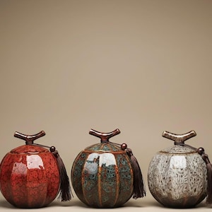 Könnte beinhalten: Drei dekorative Keramikgläser mit Deckeln und Quasten. Die Gläser sind in Rot-, Türkis- und Grautönen gehalten, jedes mit einem einzigartigen strukturierten Muster. Die Deckel haben einen astähnlichen Griff.