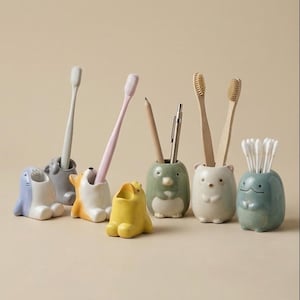 Puede incluir: Colección de soportes de cerámica con forma de animales en varios colores, cada uno con un cepillo de dientes, un bolígrafo o bastoncillos de algodón. Los soportes incluyen una foca azul, un oso gris, un corgi naranja, una rana amarilla, un pingüino verde, un oso blanco y una rana azul.
