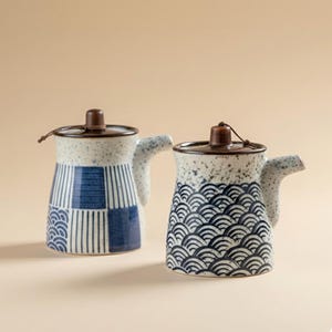 Puede incluir: Dos jarras de cerámica con tapas. Una jarra tiene un patrón a cuadros azul y blanco, y la otra tiene un patrón de olas azules. Ambas tienen un asa pequeña y una tapa de madera marrón con una cuerda. Fondo beige.