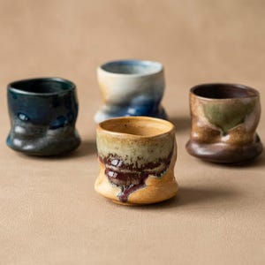 Puede incluir: Cuatro tazas de cerámica hechas a mano con formas orgánicas únicas. Las tazas presentan una variedad de colores, incluyendo azul oscuro, crema, marrón y verde, con un acabado texturizado y vidriado. Las tazas están dispuestas sobre una superficie de tono neutro.