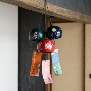 Peut inclure: Un carillon éolien suspendu composé de trois cloches en céramique arrondies de couleur vert foncé, bleu foncé et rouge, ornées de motifs floraux. Trois bandes de papier rectangulaires orange, rose et bleu clair pendent avec du texte japonais.