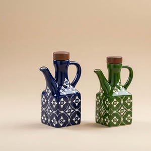 Puede incluir: Dos dispensadores de aceite de cerámica. Uno es azul marino, el otro verde. Ambos tienen una base cuadrada, un pico y un tapón de madera. Los dispensadores están decorados con un estampado floral blanco. El fondo es de color beige liso.