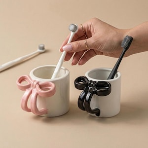 Puede incluir: Dos porta cepillos de dientes de cerámica blanca con asas en forma de lazo, uno rosa y otro negro. Cada soporte contiene un cepillo de dientes. Un tercer cepillo de dientes está en el fondo. Los soportes están sobre un fondo neutro.