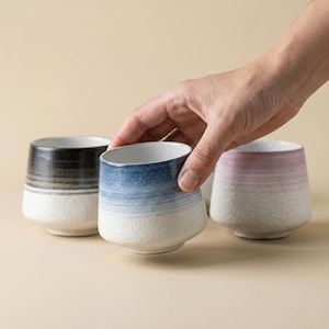 Puede incluir: Una mano sostiene una taza de cerámica azul y blanca. Se ven otras tazas negras y rosas. Las tazas tienen una superficie texturizada y un diseño moderno, perfectas para servir bebidas.