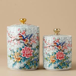 May include: Two ceramic jars with lids, decorated with a floral and bird design. The jars have a light green background with a crackle pattern. The lids are topped with gold-coloured finials. The jars are decorated with pink flowers, colourful birds, and peonies.