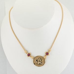 Puede incluir: Un collar de cadena de oro con un colgante redondo que presenta una escultura en relieve de dos figuras. El colgante está acentuado con cuatro pequeñas piedras preciosas rojas.