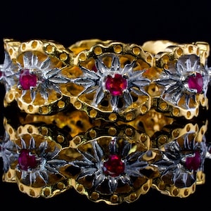 May include: A gold ring with a delicate filigree design and red gemstones. The ring is set against a black background.
