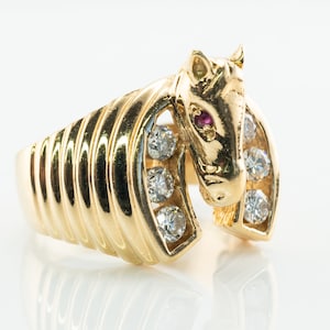 Puede incluir: Un anillo de oro con un diseño detallado de cabeza de caballo con un ojo de rubí y una herradura engastada con diamantes.
