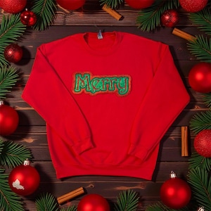May include: A red sweatshirt with the word "Merry" embroidered in green and gold. The sweatshirt is surrounded by Christmas baubles, pine branches, and cinnamon sticks, creating a festive holiday theme.