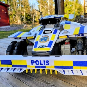 May include: A white and blue toy police car with the word "POLIS" prominently displayed. The car features black wheels, yellow and blue accents, and a Swedish coat of arms. The toy is designed with a futuristic aesthetic.