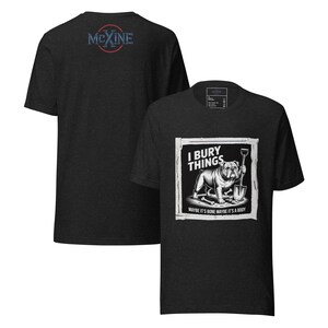Op de afbeelding: Donkergrijs t-shirt met een afbeelding van een bulldog met een schop, met de tekst "I Bury Things" en "Maybe it's bone maybe it's a body." De achterkant van het shirt heeft een cirkelvormig logo met de tekst "Mexine."